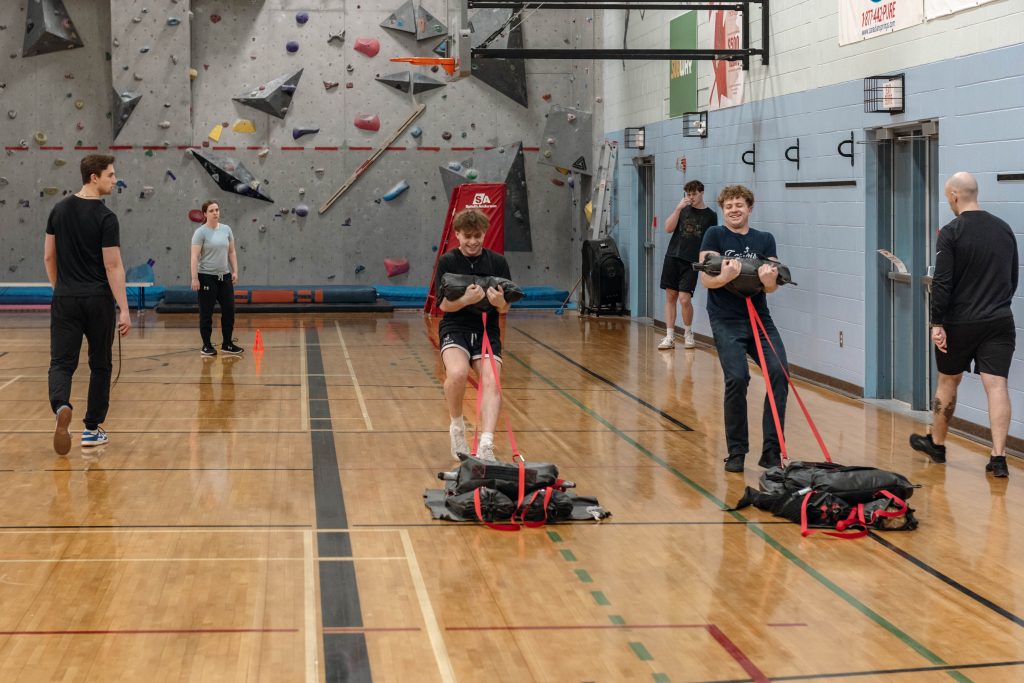 The 2025 Canadian Forces Base Halifax High School Navy Outreach Program students completing the FORCE fitness test with assistance from Personnel Support Programs Halifax fitness instructors in the Fleet Fitness and Sports Centre at His Majesty’s Canadian Dockyard. The participants are dragging black sandbags across the gym floor. / Les élèves du Programme de sensibilisation de la Marine dans les écoles secondaires 2025 passent le test FORCE avec l’aide des instructeurs de conditionnement physique des Programmes de soutien au personnel au Centre de conditionnement physique et de sports de la Flotte à l’arsenal de Sa Majesté. Les participants traînent des sacs de sable noirs sur le sol du gymnase.