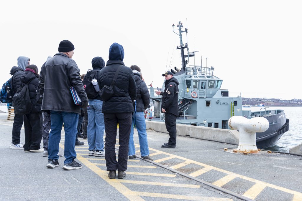 A group of 2025 CFB Halifax High School Navy Outreach Program students standing in His Majesty’s Canadian Dockyard next to a grey King’s Harbour Master tugboat afloat in Halifax Harbour. / Un groupe d’élèves participant au Programme de sensibilisation de la Marine dans les écoles secondaires 2025 à la BFC Halifax se tient à l’arsenal canadien de Sa Majesté à côté d’un grand remorqueur du capitaine du port de Sa Majesté à flot dans le port d’Halifax.