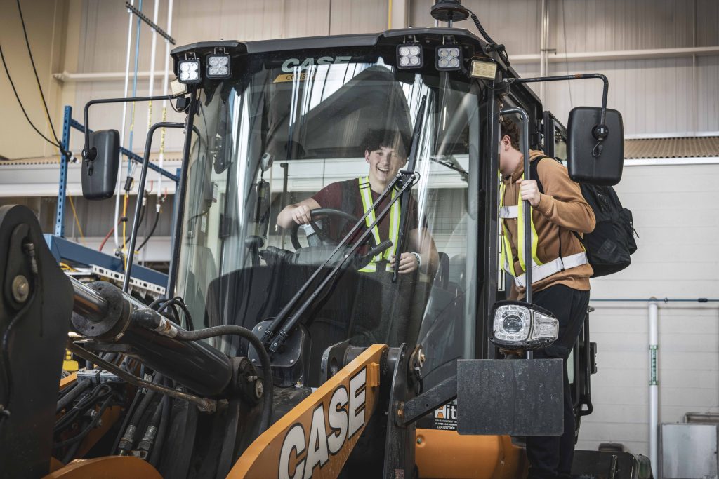 Two 2025 CFB Halifax High School Navy Outreach Program students exploring a heavy equipment vehicle at the Base Logistics Transport, Electrical, and Mechanical Engineering Division headquarters. One student is sitting in the driver’s seat, and one is standing in the doorway of the vehicle. / Deux élèves participant au Programme de sensibilisation de la Marine dans les écoles secondaires 2025 à la BFC Halifax inspectent un véhicule lourd au quartier général de la Division du transport et du génie électrique et mécanique du Service de la logistique de la base. Un élève est assis au volant et un autre se tient à la porte du véhicule.